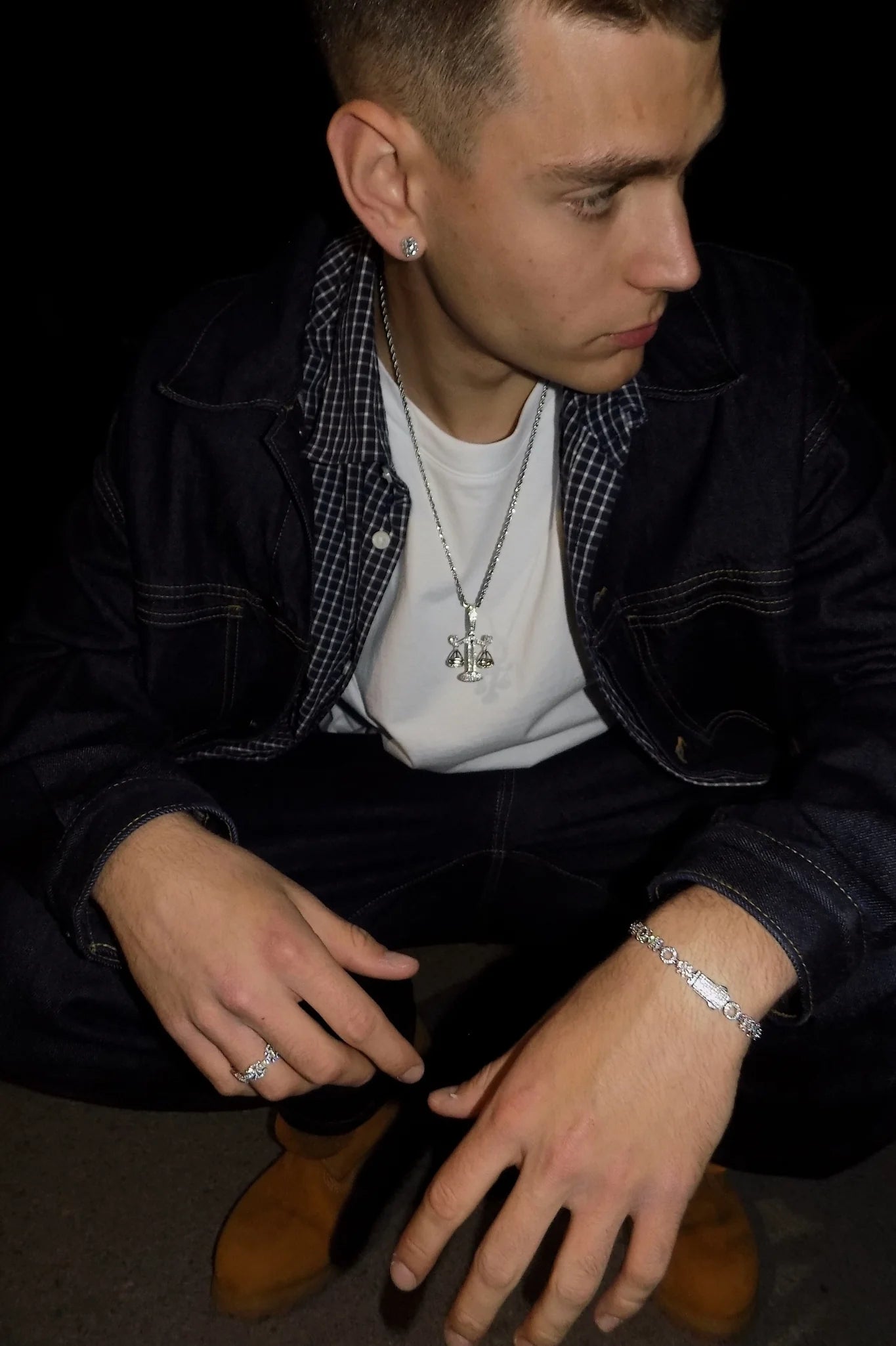 Crouching model in a dark denim outfit wearing the ÉLIX 925 silver O-X bracelet with pavé moissanite stones on the right wrist — AETHER