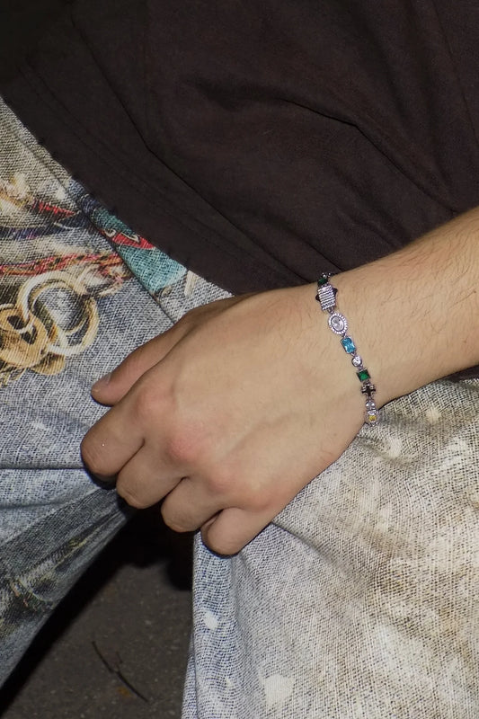 Close-up of a person’s wrist wearing the ÓPALLION 925 silver bracelet with multicolored stones and enamel cross motifs, resting on patterned denim — AETHER